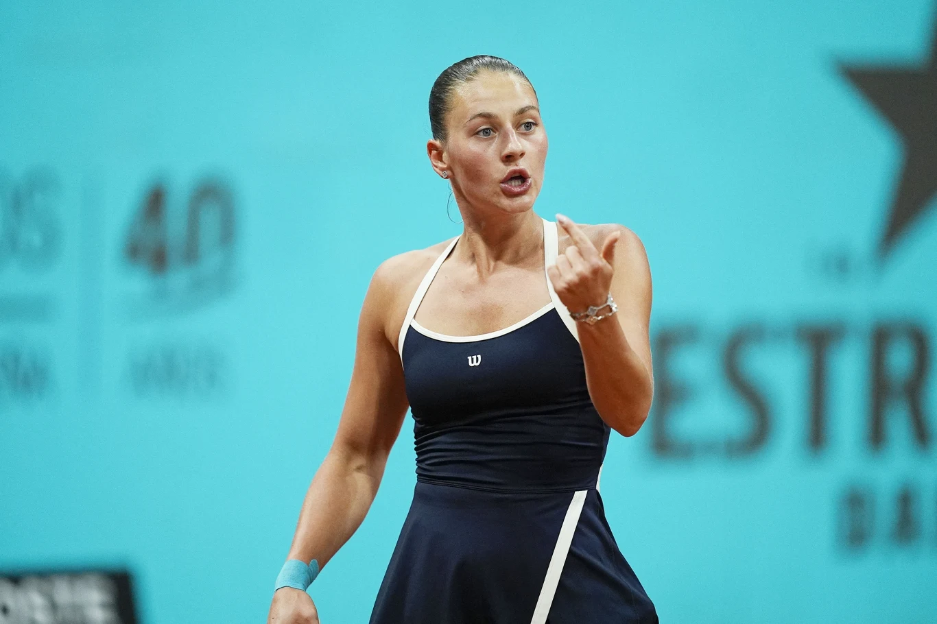 Marta Kostiuk Tenisistka na korcie w sportowym stroju, wykonująca gest ręką z wyrazem emocji na twarzy, na tle niebieskiego baneru turniejowego.