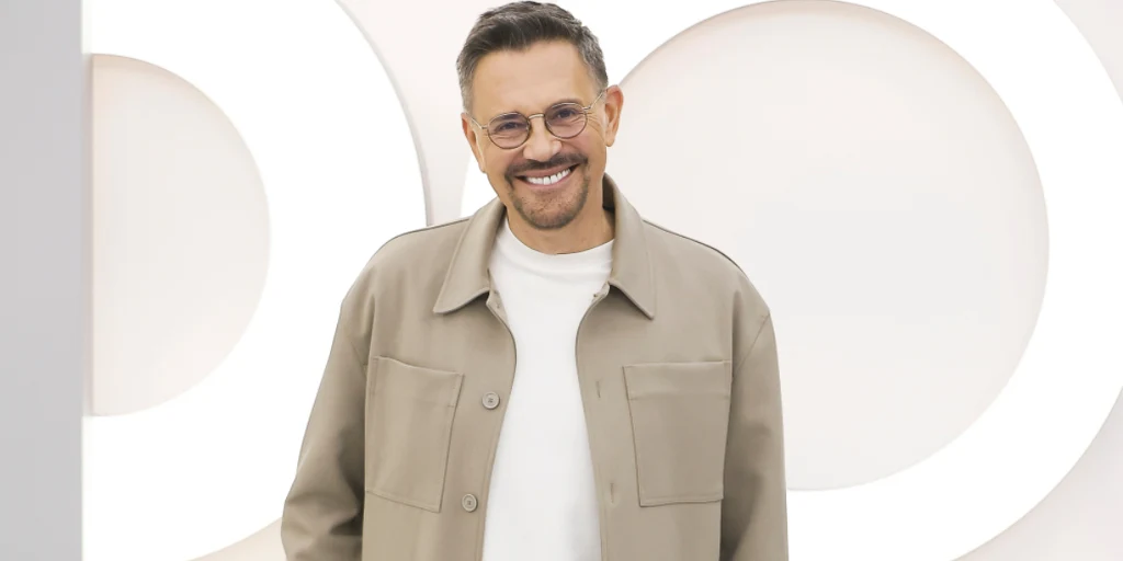 Uśmiechnięty mężczyzna w okularach, jasnej kurtce i białej koszulce stoi na tle białej ściany z geometrycznymi okręgami podświetlonymi światłem.