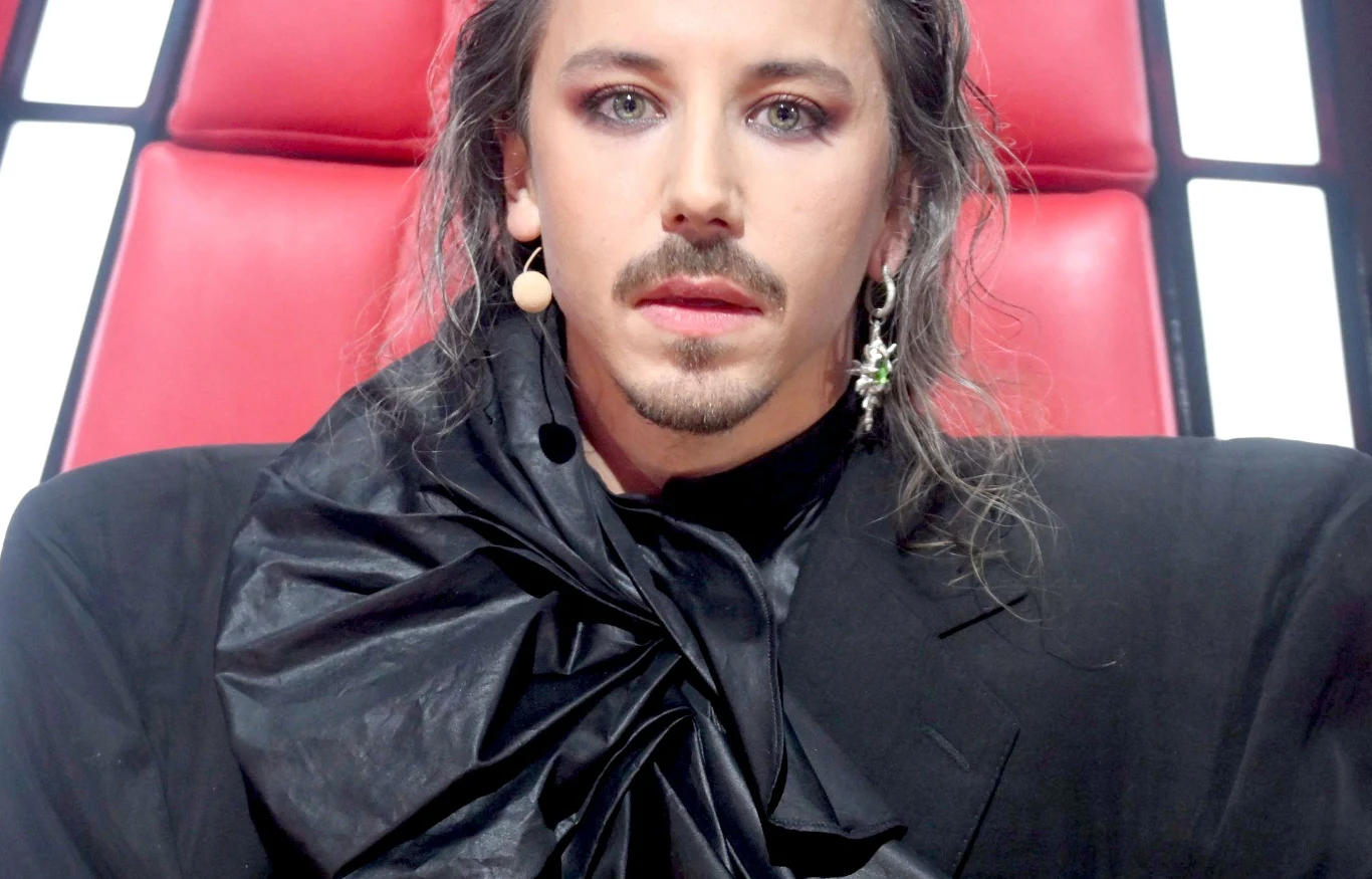 Osoba siedząca na czerwonym fotelu w eleganckim, czarnym stroju ozdobionym efektowną falbaną, z długimi włosami, makijażem i charakterystyczną kolczykiem w uchu, widoczna na tle kontrastujących jasnych paneli.