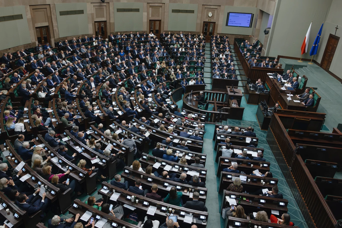 Najnowszy sondaż. Cztery partie w Sejmie, KO powiększa przewagę Sala pełna posłów podczas obrad parlamentu, uczestnicy siedzą w ławach ustawionych półkoliście, na mównicy osoby prowadzące obrady, po prawej stronie flagi Polski i Unii Europejskiej.