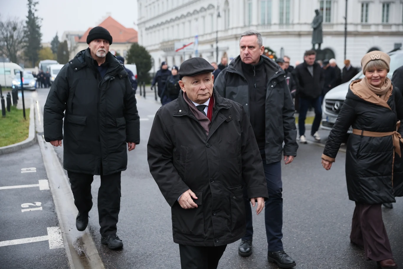 Jarosław Kaczyński na miesięcznicy smoleńskiej Grupa osób idących ulicą w centrum miasta, na czele starszy mężczyzna w ciemnym płaszczu i kapeluszu, w towarzystwie ochroniarzy i kobiety uśmiechającej się, w tle budynek rządowy oraz samochód.