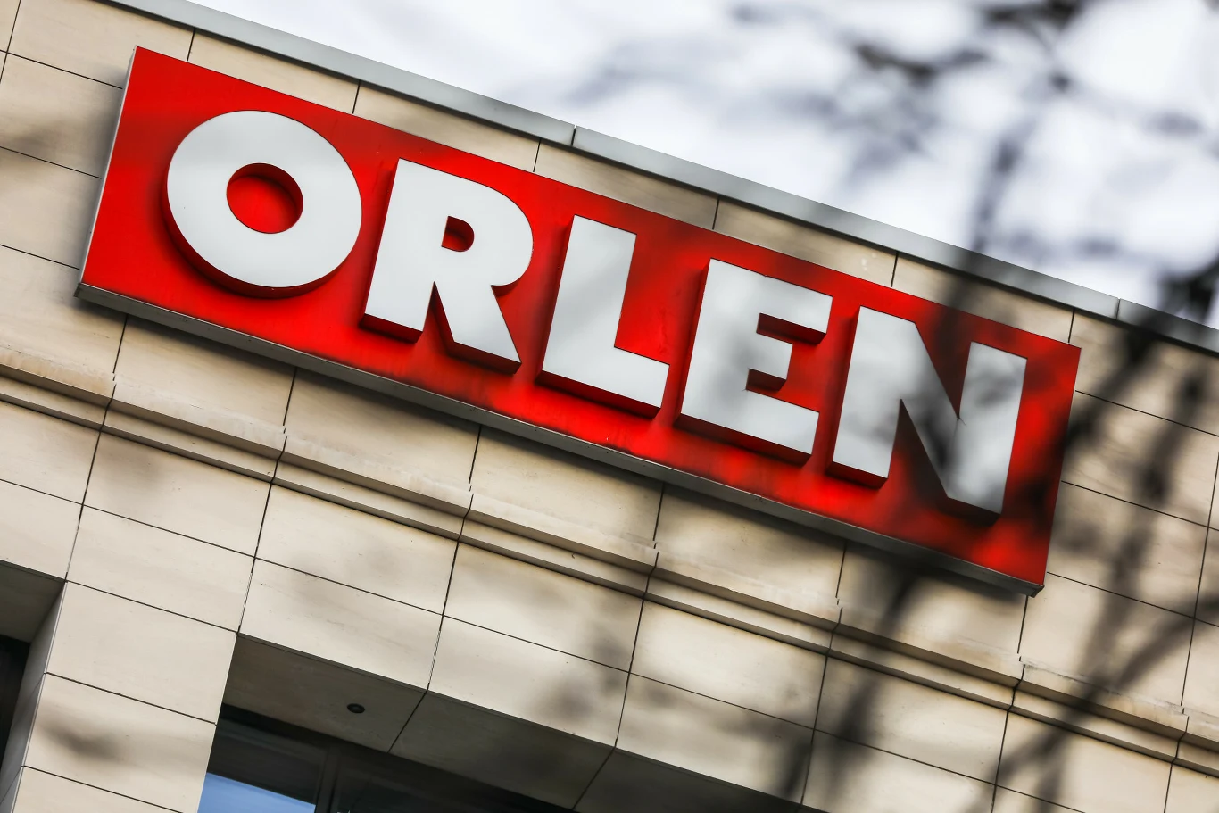 Logo firmy Orlen na czerwonym tle umieszczone na elewacji nowoczesnego budynku. Białe, duże litery wyraźnie wyróżniają się na tle jasnej ściany. W kadrze widoczne są również delikatne cienie gałęzi.