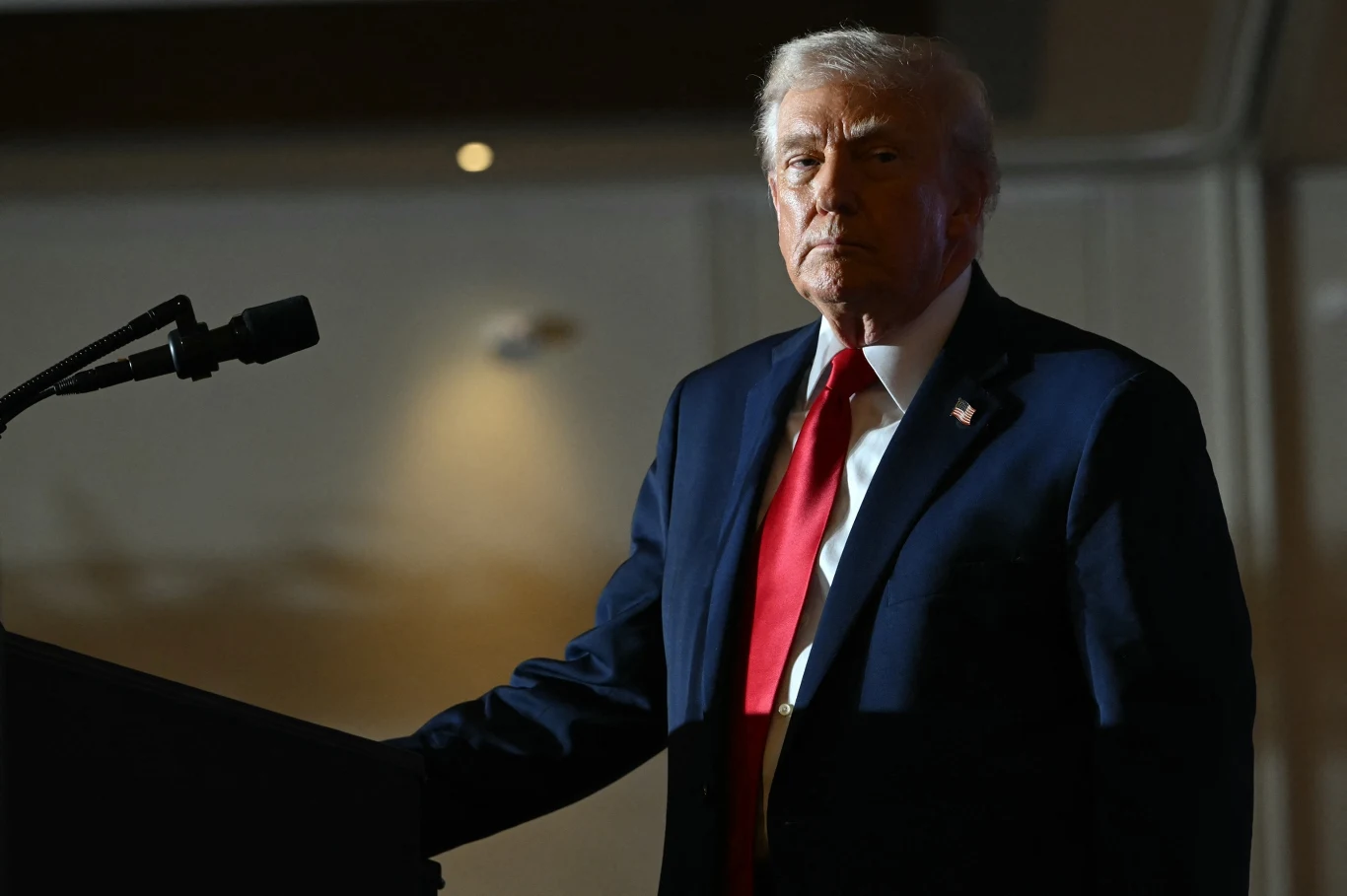 Starszy mężczyzna w ciemnym garniturze i czerwonym krawacie stoi przy mikrofonie podświetlony punktowym światłem, reszta sceny jest zacieniona, tło neutralne, nastrojowe oświetlenie podkreśla powagę sytuacji.