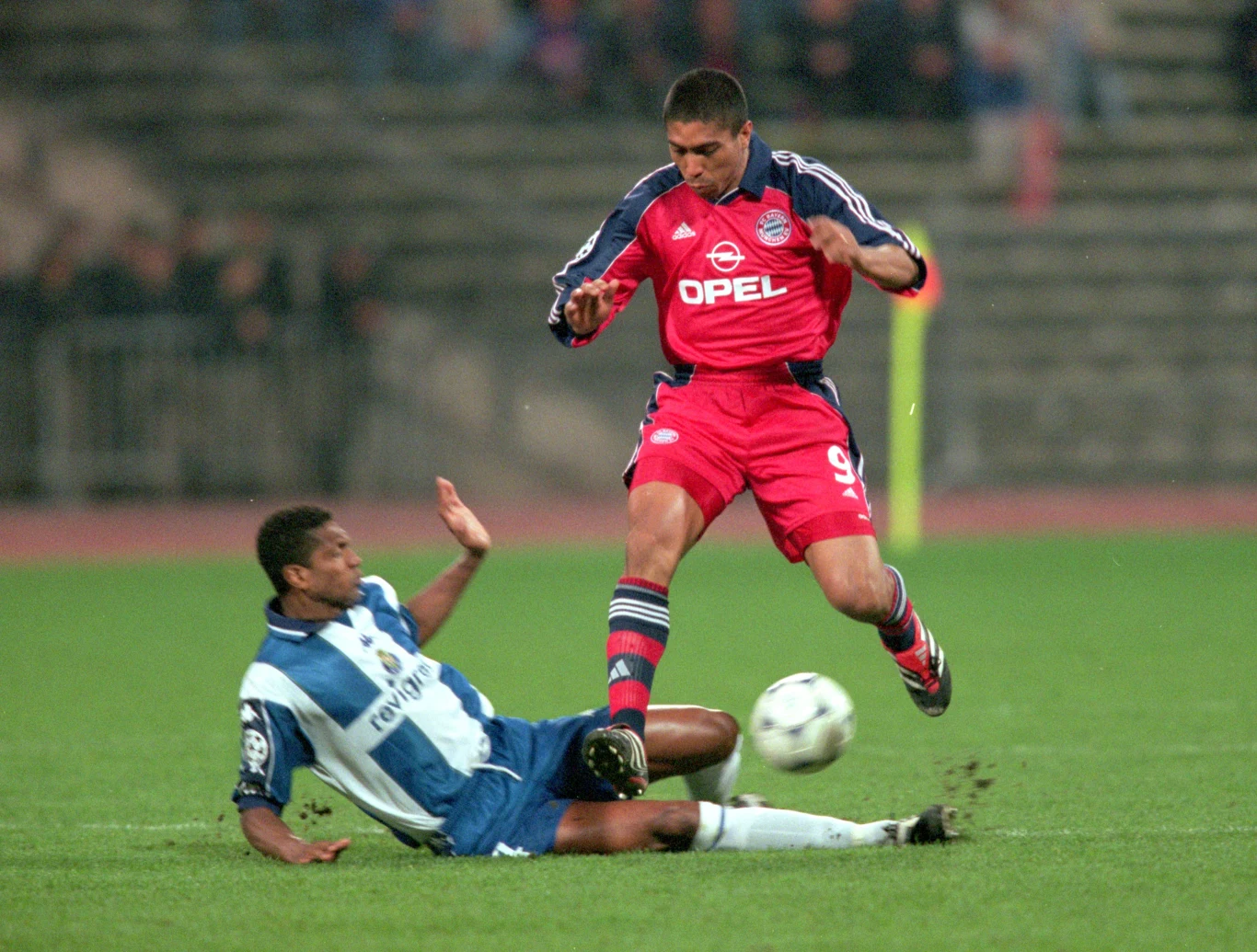 Dwóch piłkarzy podczas dynamicznej akcji na boisku, jeden z nich próbuje odebrać piłkę przeciwnikowi wślizgiem. Tło stanowi murawa stadionu oraz zamazane trybuny z widownią.