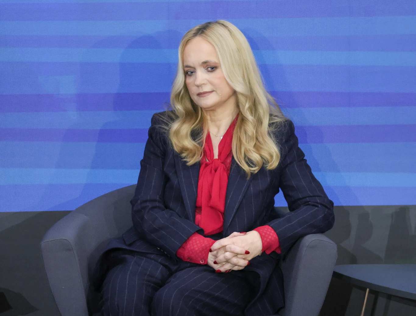 Minister Sobierańska-Grenda chce uzdrowić finanse NFZ Kobieta o blond włosach ubrana w granatowy garnitur w prążki i czerwoną bluzkę siedzi na fotelu na tle niebieskiej ściany.