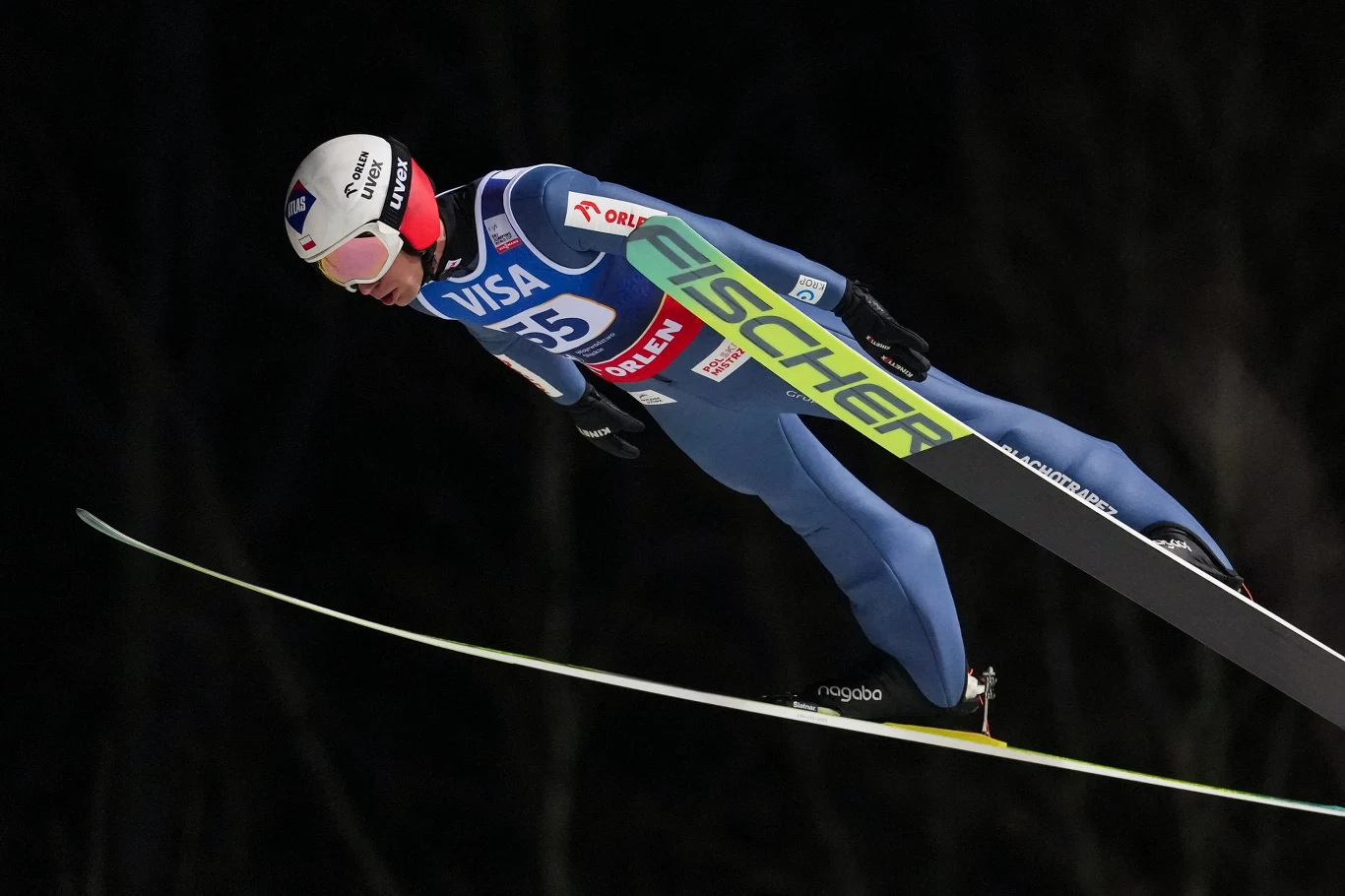 Sportowiec w niebieskim kombinezonie i kasku wykonuje skok narciarski w locie z nartą marki Fischer na tle ciemnego nieba.