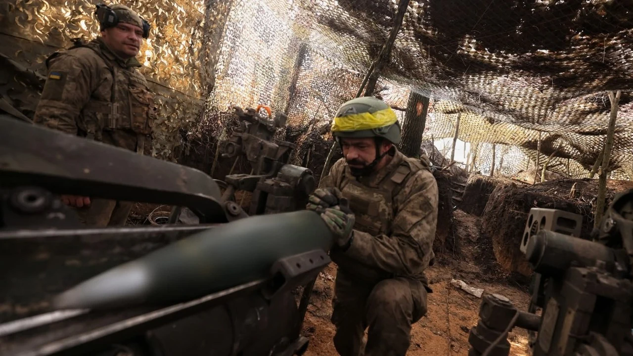 Spada tempo pomocy wojskowej dla Ukrainy Żołnierz przygotowuje pocisk artyleryjski do załadowania do działa w okopanym stanowisku, wokół widoczna jest siatka maskująca i sprzęt wojskowy, w tle inny żołnierz obserwuje sytuację.
