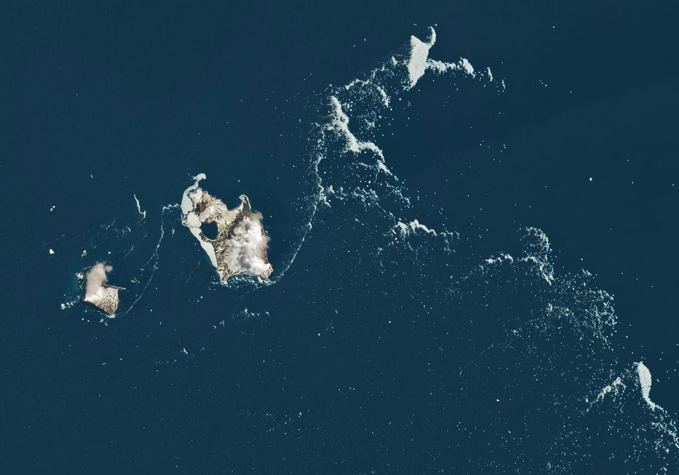 Grupa niewielkich wysp otoczona ciemnoniebieskim oceanem, ostre linie brzegowe wyraźnie kontrastują z wodą, wokół wysp widoczne ślady piany morskiej.