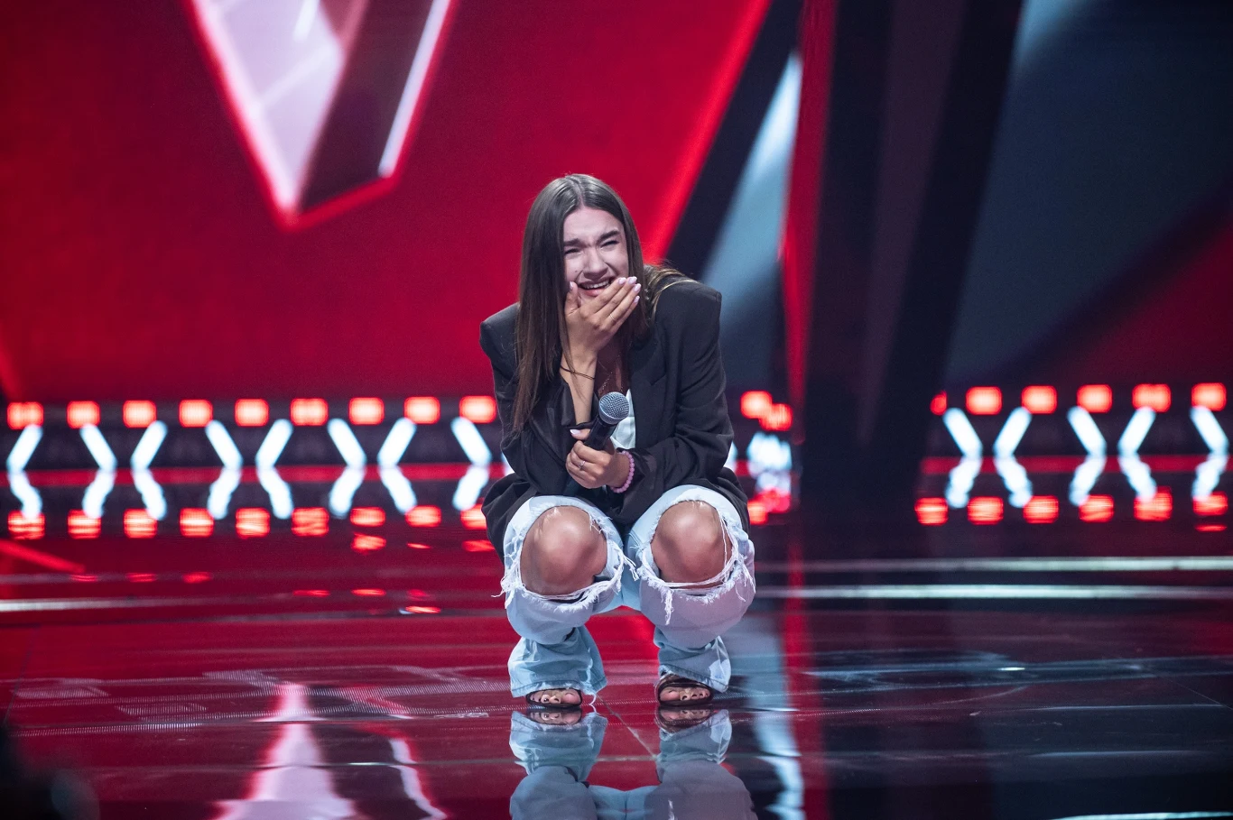 Julia Wasielewska dotarła do ćwierćfinału 16. edycji "The Voice of Poland" Kobieta z długimi włosami klęczy na scenie z mikrofonem w dłoni, uśmiecha się szeroko i jest wyraźnie wzruszona, ubrana w podarte jeansy oraz czarną marynarkę, w tle charakterystyczna scenografia programu talent show z czerwonymi i czarnymi elementami ...