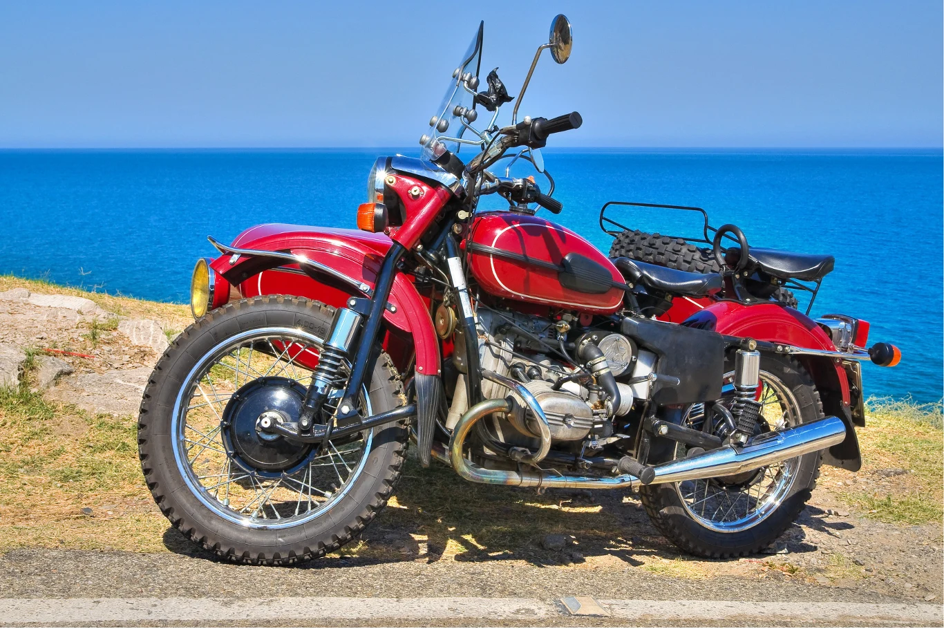 Ural w tarapatach. Szykuje się ważna zmiana w gamie modelowej. Czerwony zabytkowy motocykl z wózkiem bocznym stoi zaparkowany na poboczu, w tle rozciąga się błękitne morze i czyste niebo.
