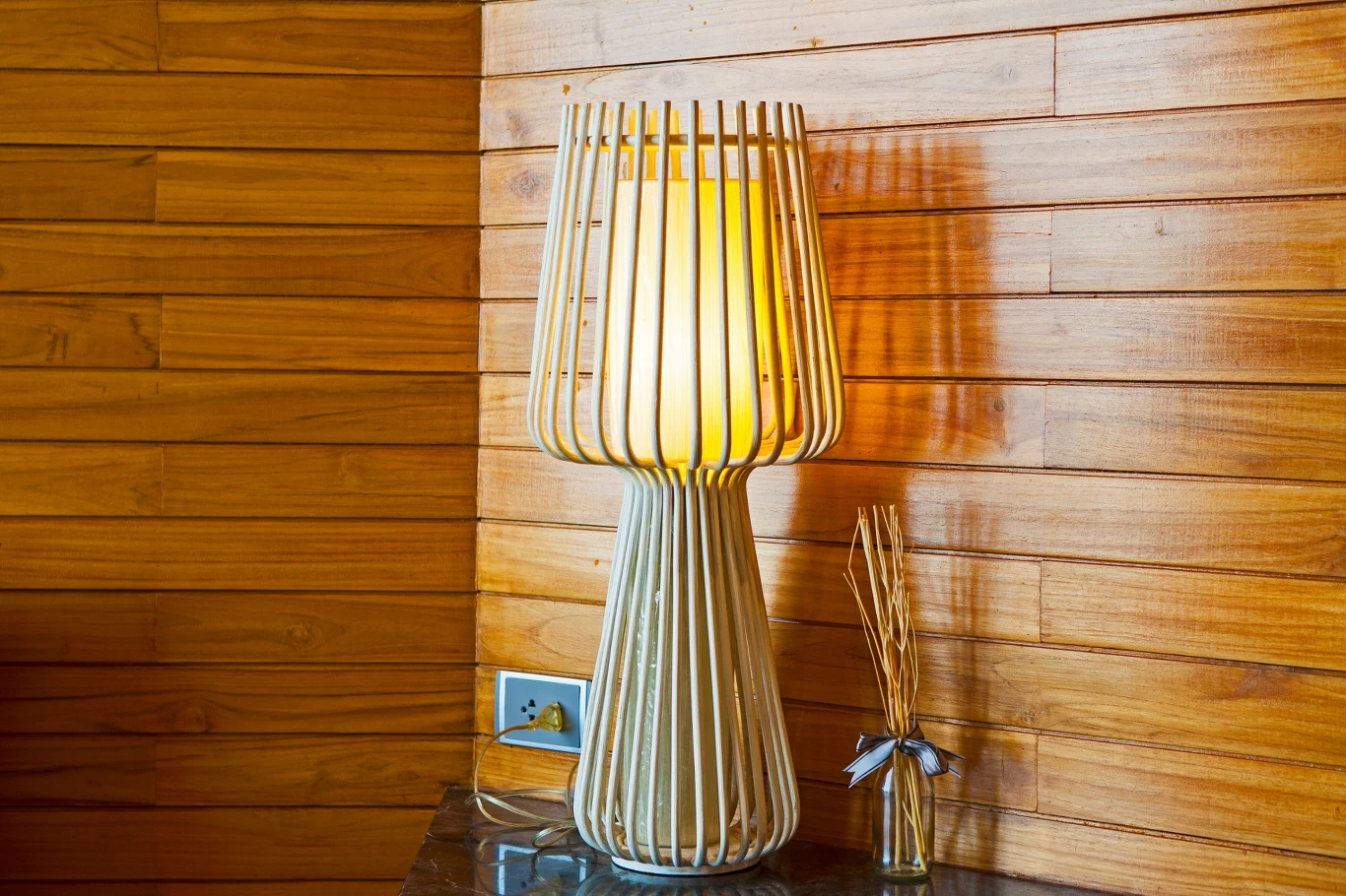 Stolik z lampą o oryginalnym, ażurowym kloszu emitującą ciepłe światło, obok dekoracyjne patyczki zapachowe, a całość ustawiona na tle drewnianej ściany.