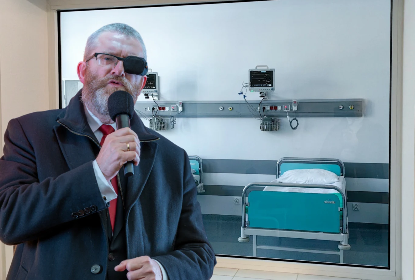 Grzegorz Braun interweniował w oleśnickim szpitalu. Prokuratura umorzyła śledztwo dotyczące lekarki Gizeli Jagielskiej Mężczyzna z opaską na oku i mikrofonem w dłoni stoi na tle szpitalnej sali z pustymi łóżkami i sprzętem medycznym pod ścianą.