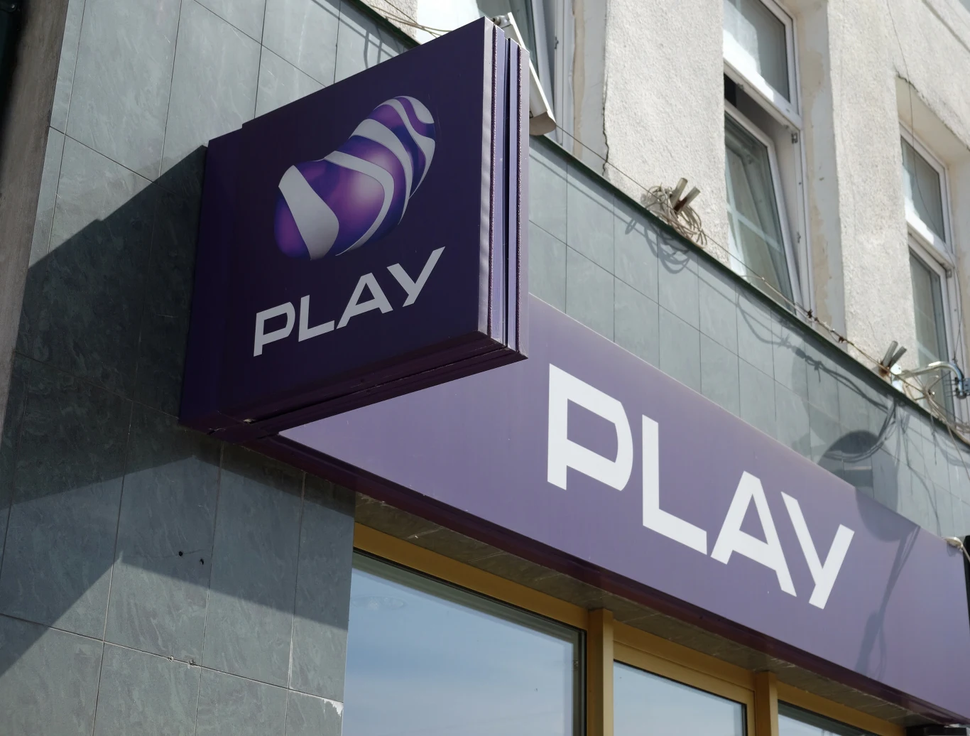UOKiK ukarał Play karą (zdj. ilustracyjne) Szyld sklepu sieci telekomunikacyjnej Play z logo w odcieniach fioletu zamontowany na elewacji budynku obok okien.