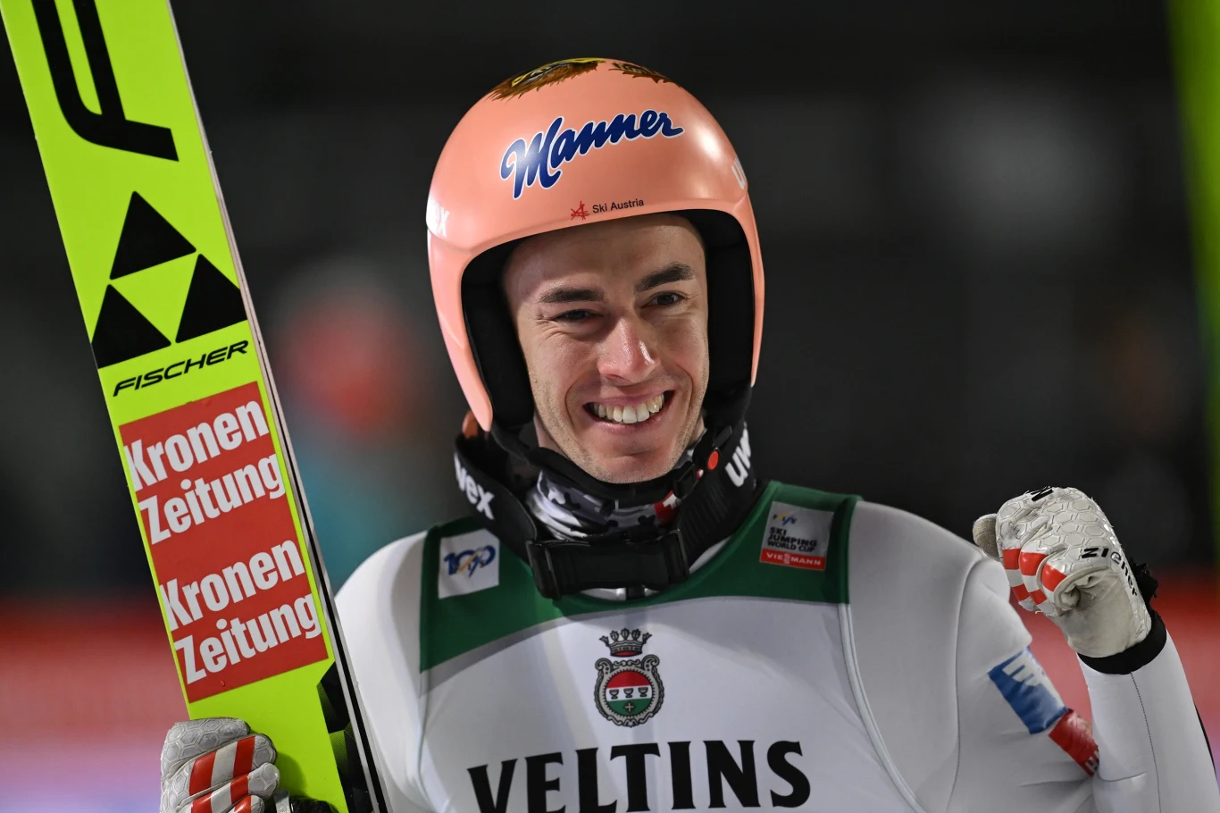 Stefan Kraft Zawodnik skoków narciarskich w sportowym stroju i kasku, z uśmiechem na twarzy, trzymający narty na tle ciemnego, rozmazanego tła ze sztucznym oświetleniem.