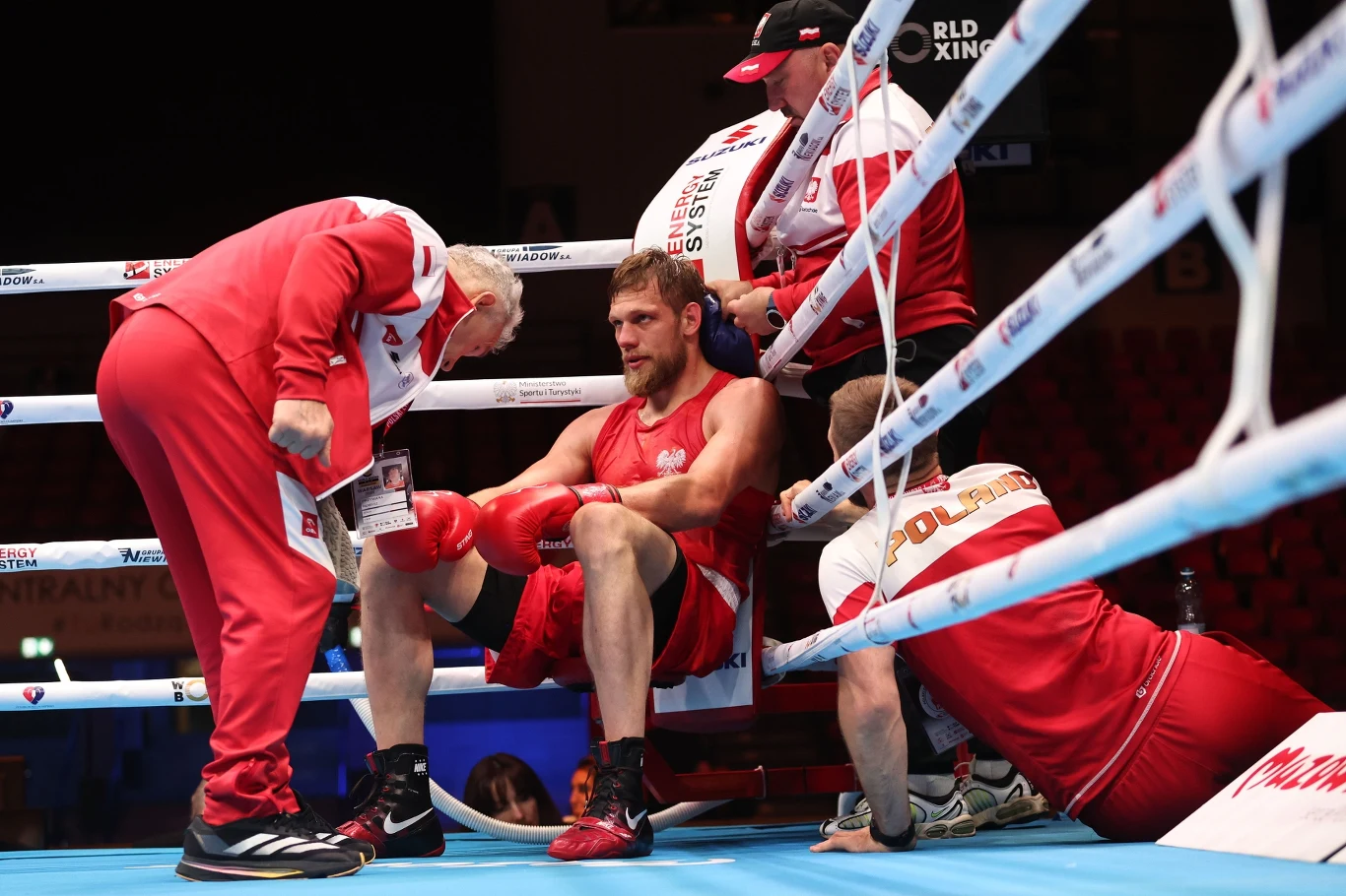Aleksiej Sewostjanow w narożniku Bokser siedzący na ringu w narożniku otoczony przez trzech członków sztabu szkoleniowego. Wszyscy ubrani w czerwono-białe stroje. Bokser wygląda na zmęczonego, sztab udziela mu wskazówek i wsparcia.