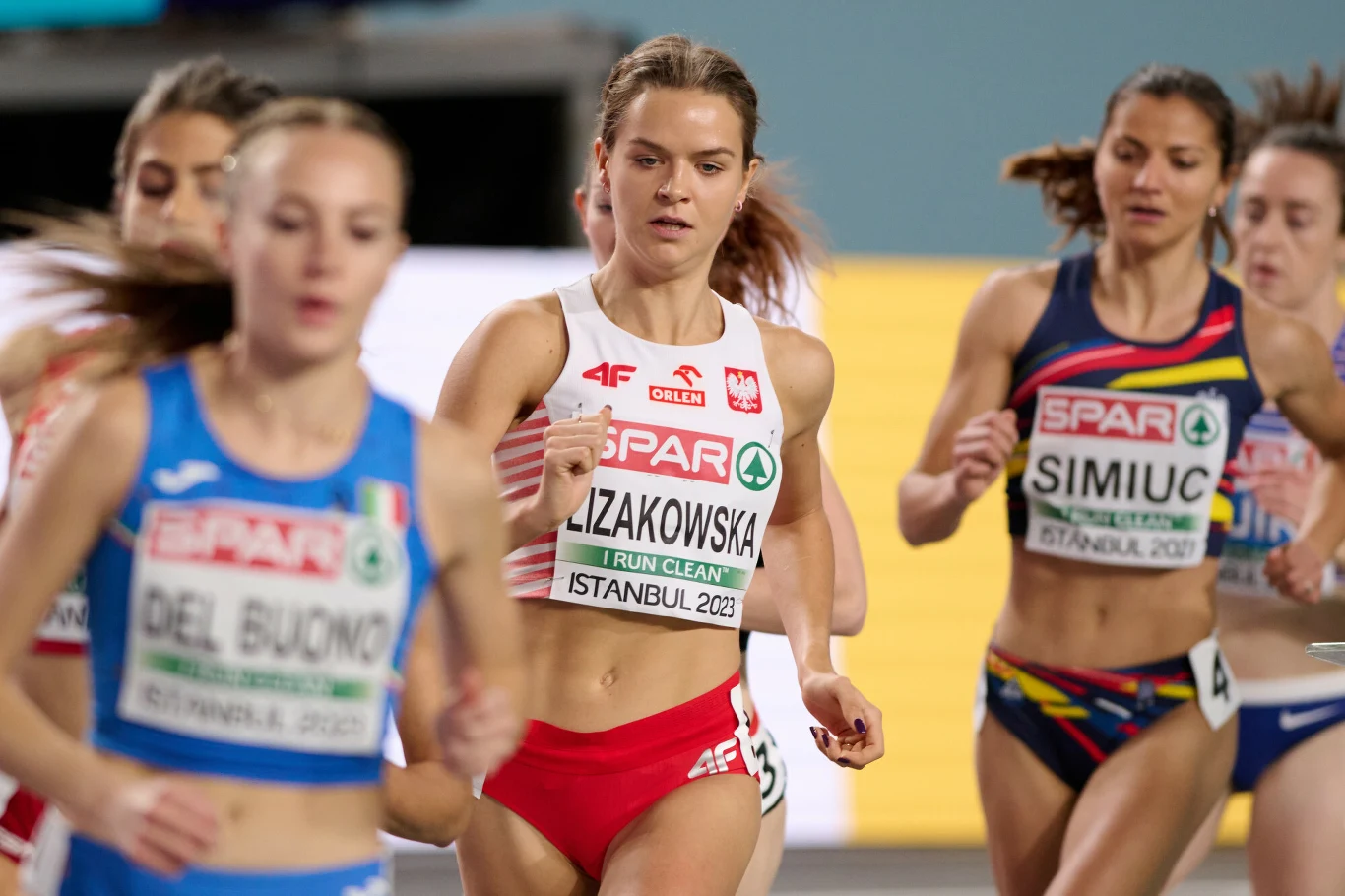 Zawodniczki biorące udział w biegu lekkoatletycznym podczas zawodów. Centralnie widoczna polska biegaczka w biało-czerwonym stroju, wokół niej inne zawodniczki z różnych krajów. Panuje sportowa rywalizacja, a stroje posiadają identyfikatory i oznaczeni...