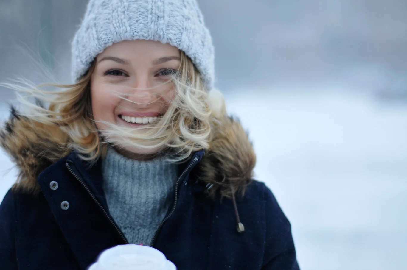 Numerologiczne przewidywania na święta: kto będzie miał najlepsze dni Uśmiechnięta kobieta w zimowej kurtce z futrzanym kołnierzem, nosząca wełnianą czapkę, trzyma kubek gorącego napoju, w tle śnieżny krajobraz.