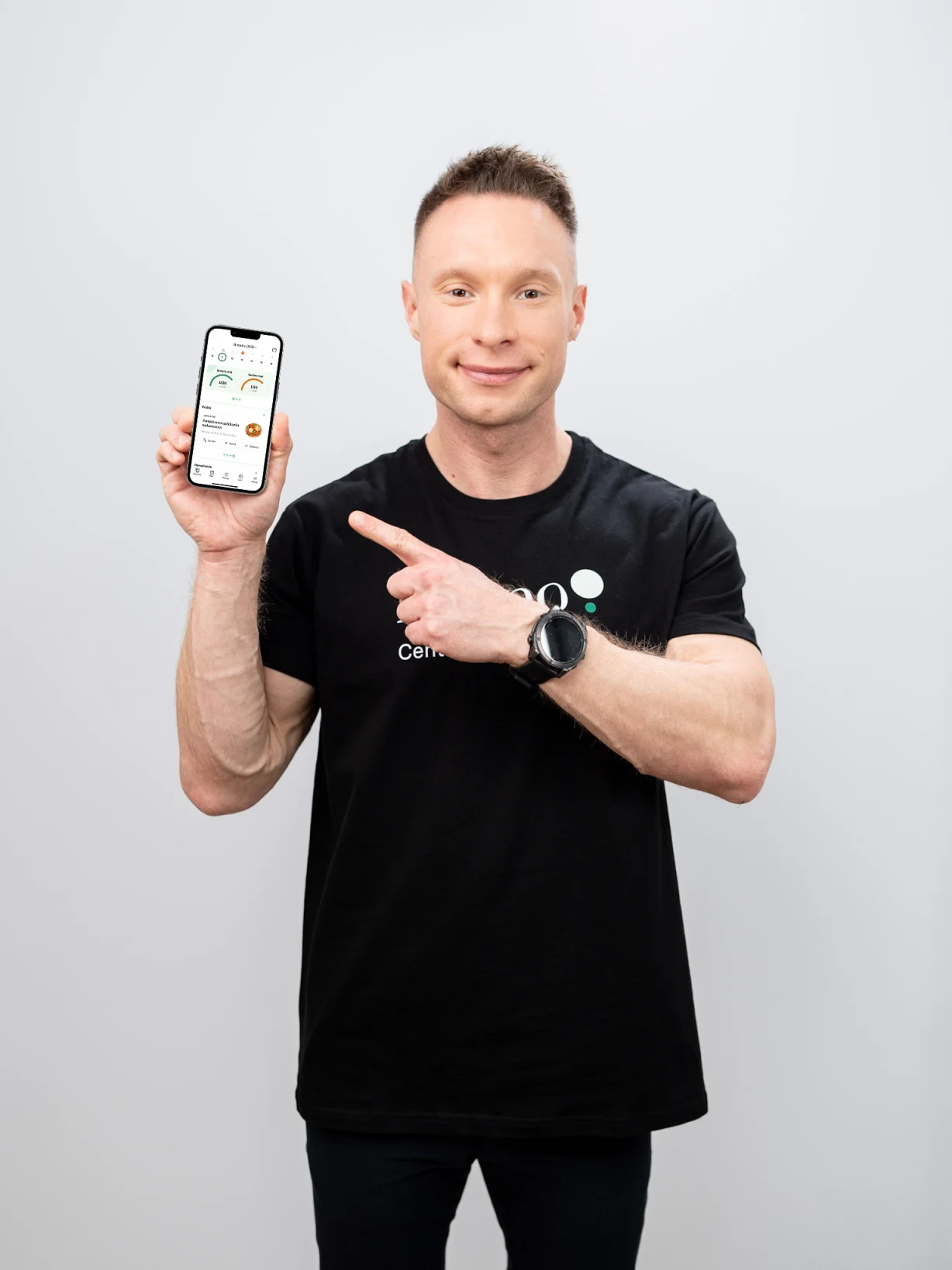 Mężczyzna w czarnej koszulce z zegarkiem na nadgarstku trzyma w jednej ręce smartfon z włączonym ekranem i drugą ręką wskazuje na telefon. Jasne, neutralne tło i uśmiech na twarzy podkreślają pozytywny charakter sceny.