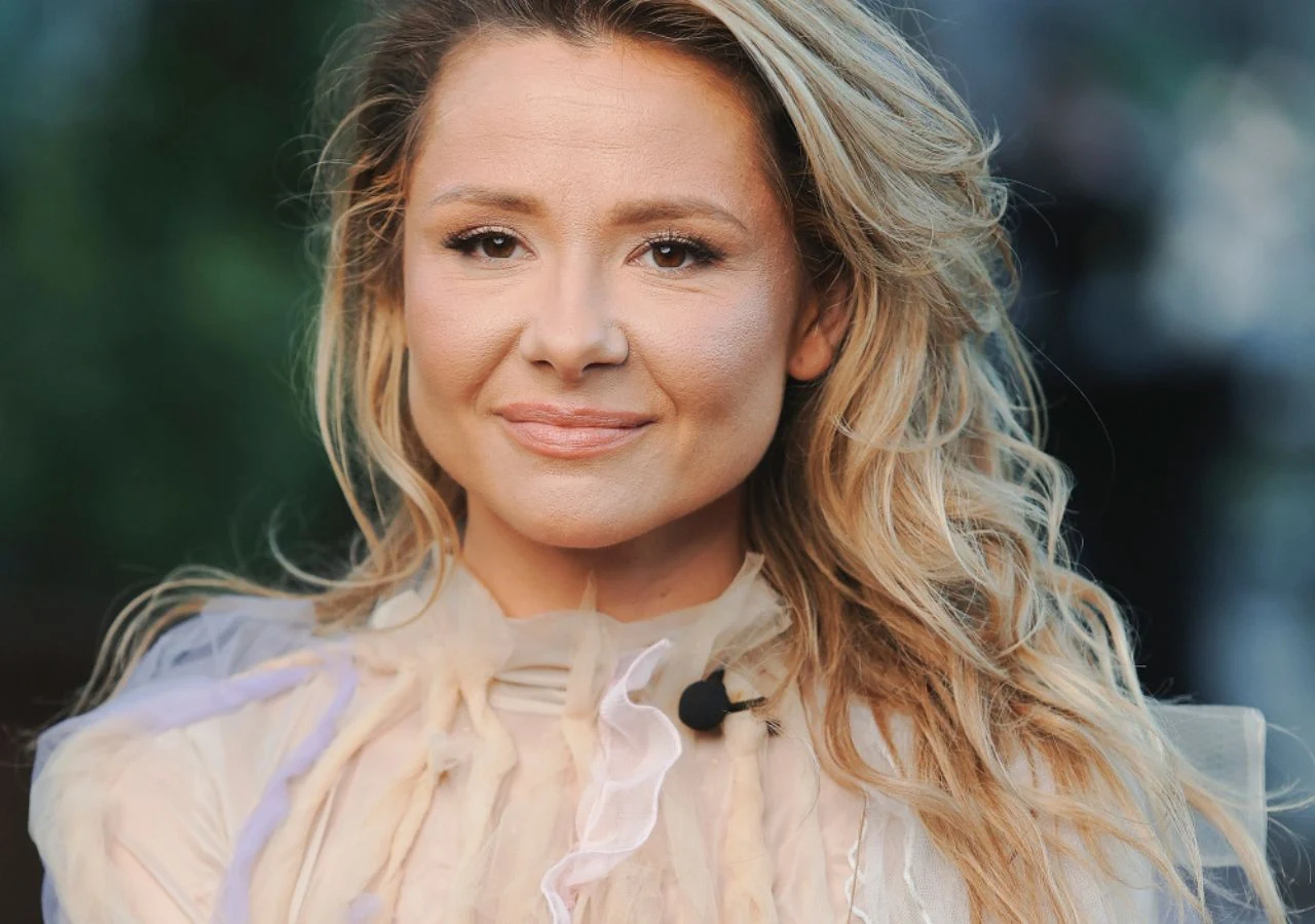 Anna Karwan Uśmiechnięta kobieta o blond włosach, ubrana w jasną, elegancką bluzkę, patrzy prosto w obiektyw. Na ramieniu widoczny mikrofon przypinany. Tło rozmyte, skupiające uwagę na twarzy.