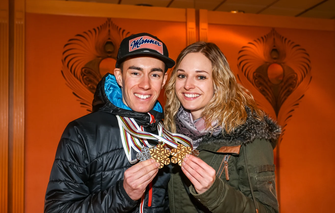 Stefan Kraft z partnerką Marisą Uśmiechnięta para młodych ludzi pozująca razem, trzymając w dłoniach kilka medali sportowych na tle ozdobnej ściany.