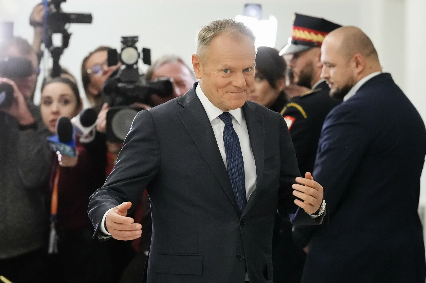 Koalicja Obywatelska premiera Donalda Tuska umacnia się na pozycji lidera w nowym sondażu Mężczyzna w garniturze i krawacie uśmiecha się, zbliżając się w kierunku aparatu, w tle grupa dziennikarzy oraz osoby w mundurach, wielu fotografów kieruje obiektywy na centralną postać.