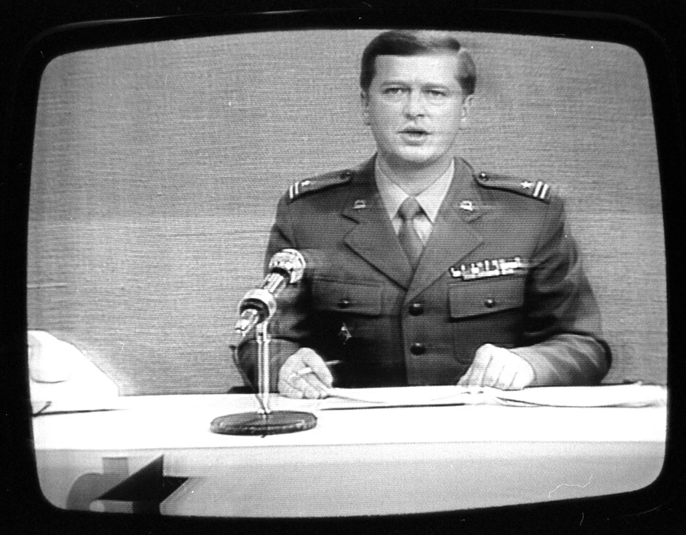 14 grudnia 1981 r. Major WP w Dzienniku Telewizyjnym Mężczyzna w mundurze wojskowym siedzi za biurkiem, mówi do mikrofonu, w tle jednolite tło, całość widoczna przez ekran telewizora.