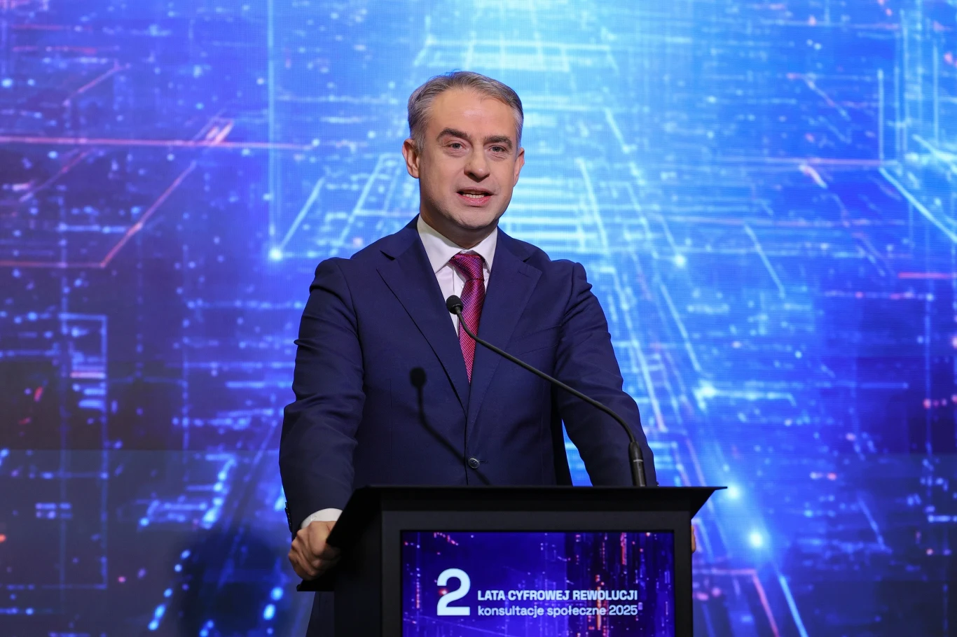 Wicepremier, minister cyfryzacji Krzysztof Gawkowski podczas konferencji prasowej po inauguracji konsultacji społecznych Ministerstwa Cyfryzacji "2 lata cyfrowej rewolucji" Mężczyzna w garniturze i pod krawatem przemawia przy mównicy na tle efektownej, cyfrowej grafiki ze schematycznymi liniami i światłami. Na mównicy znajduje się napis informujący o drugiej rocznicy cyfrowej rewolucji i konsultacjach społecznych.