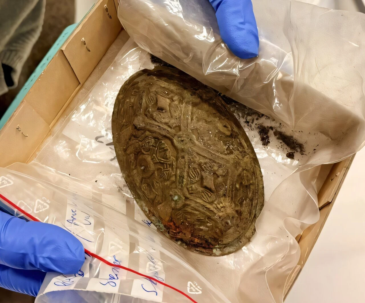 Ozdoby znalezione w grobie z czasów wikingów przykuły uwagę naukowców. Owalny artefakt pokryty wzorami znajduje się w plastikowej torbie ochronnej, trzymanej przez osobę w niebieskich rękawiczkach, umieszczony na kartonowej podkładce w laboratorium.