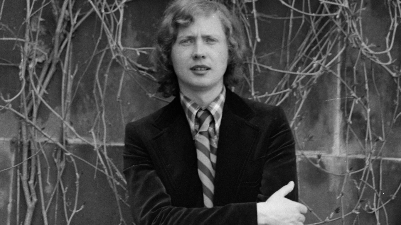 Marek Grechuta (1960) Mężczyzna w średnim wieku z kręconymi włosami w marynarce i krawacie stoi na tle ściany porośniętej suchymi pnączami, jego ręce są skrzyżowane.