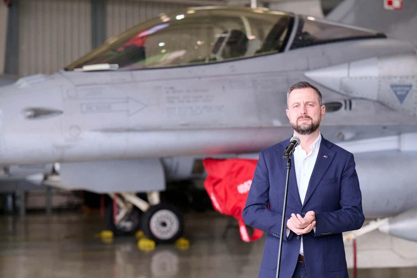 Władysław Kosiniak-Kamysz zabrał głos ws. ewentualnego przekazania Ukrainie MiG-29 (zdj. ilustracyjne) Mężczyzna ubrany w garnitur stojący przed mikrofonem w hangarze lotniczym, w tle widoczny myśliwiec wojskowy F-16 z polskimi znakami rozpoznawczymi, elementy wyposażenia technicznego przy samolocie.