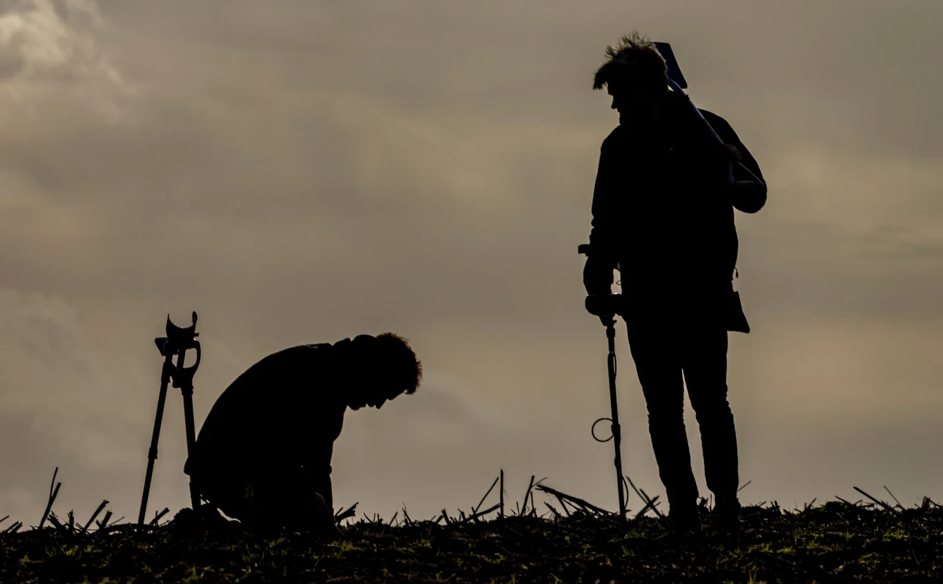 Podczas przeszukiwania terenu z wykrywaczem metali doszło do fascynującego odkrycia, które stało się w Norwegii sensacją. Zdj. ilustracyjne. Dwie osoby w ciemnych sylwetkach, jedna stoi z wykrywaczem metalu, druga jest pochylona nad ziemią, obok nich widoczny jest statyw. Na pierwszym planie fragment pola z suchą roślinnością, tło w chłodnych barwach podkreślone zachmurzeniem nieba.