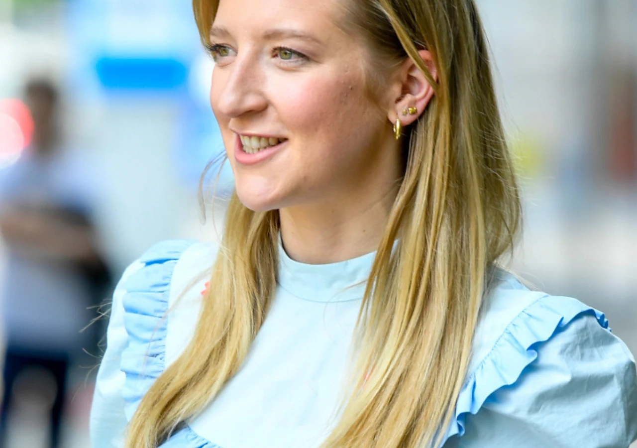 Lara Gessler Uśmiechnięta młoda kobieta o długich blond włosach, ubrana w jasnoniebieską bluzkę z falbanami, na tle lekko rozmytego miejskiego krajobrazu.