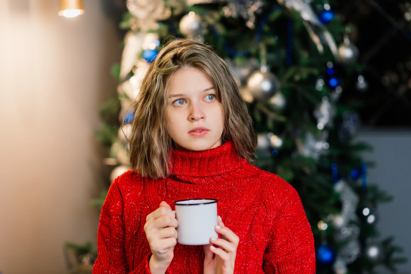 Młoda kobieta w czerwonym swetrze trzyma białą filiżankę i patrzy w bok, w tle widoczne ozdobione choinka i świąteczne dekoracje.