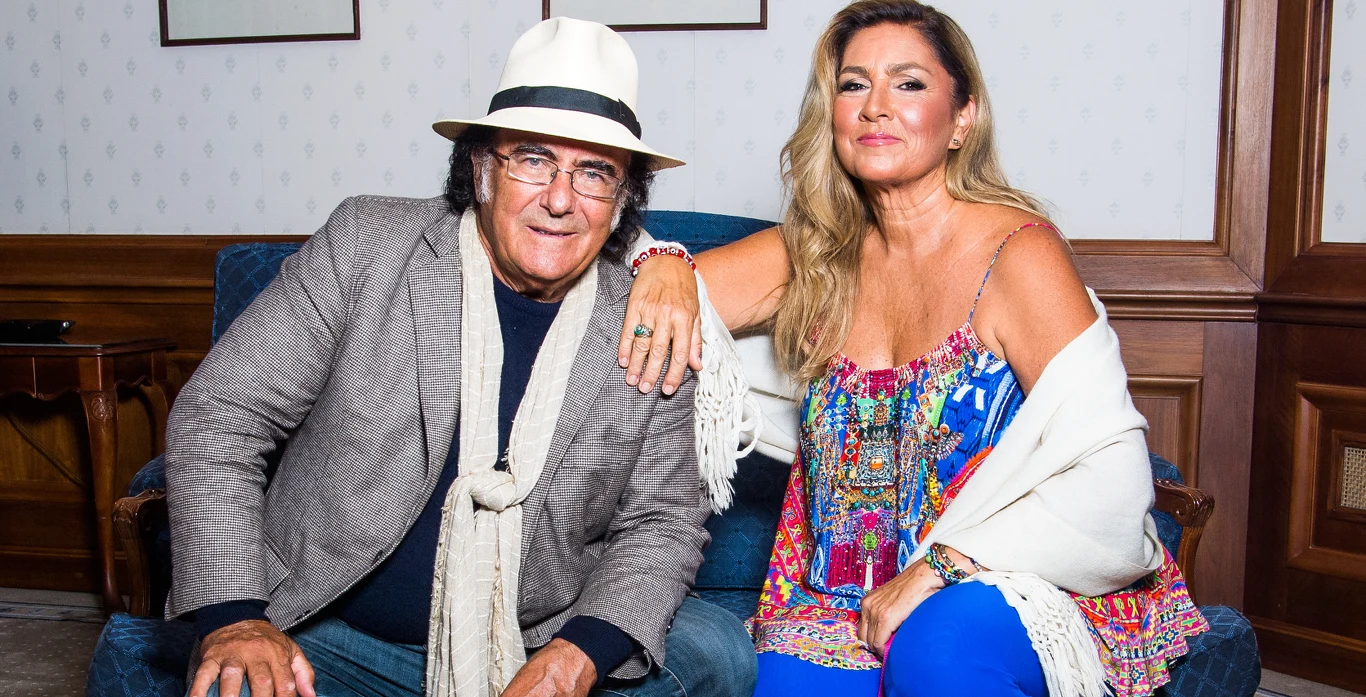 Al Bano i Romina Power nadal utrzymują kontakt Dwie elegancko ubrane osoby siedzą na niebieskiej sofie w stylowym pomieszczeniu, kobieta w kolorowej sukience i szalu opiera rękę na ramieniu mężczyzny w kapeluszu, marynarce i szalu.