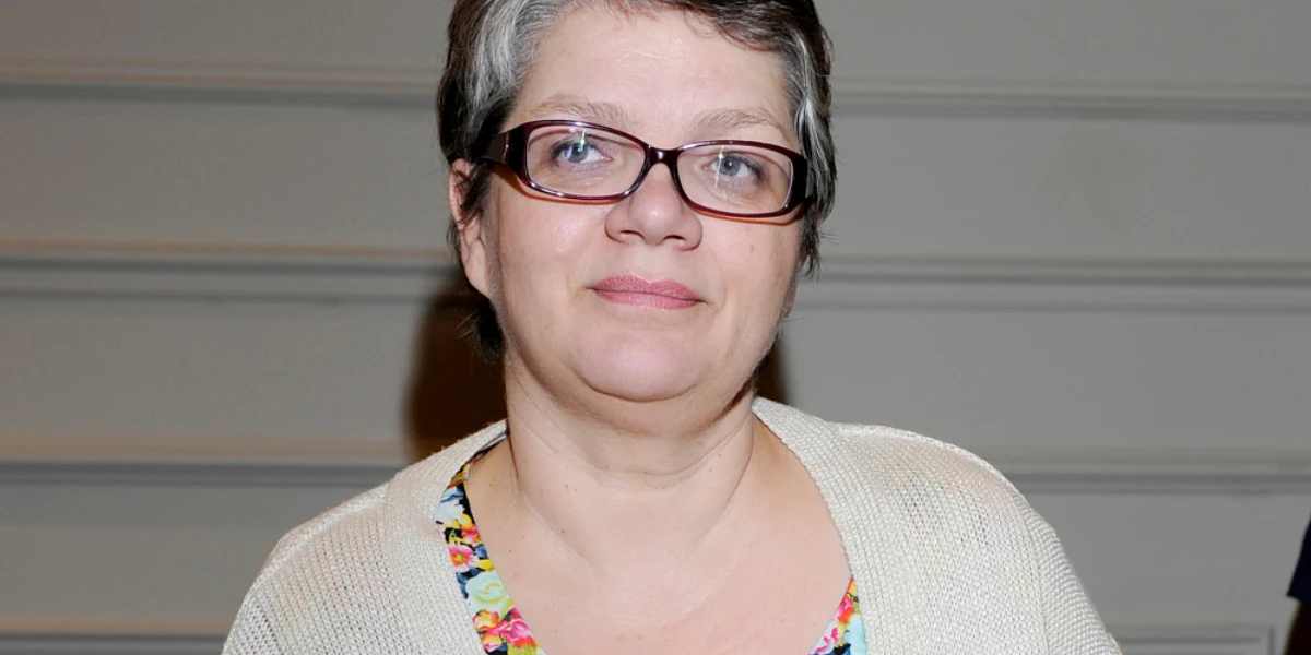 Dorota Zawadzka Kobieta w okularach z krótkimi, szpakowatymi włosami, uśmiechnięta, ubrana w jasny sweter i wzorzystą bluzkę, siedzi na tle jasnej ściany.