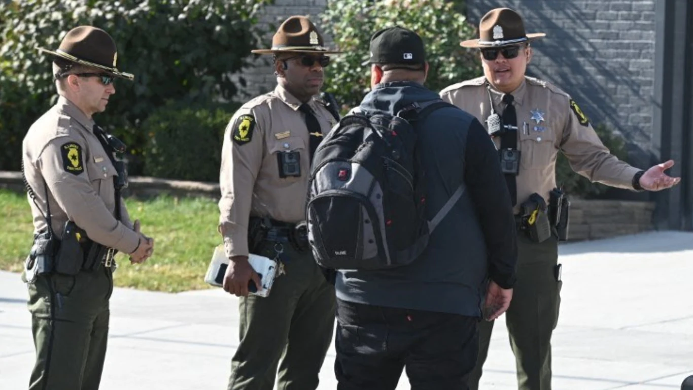 Deportacje Polaków z USA. Nasila się presja amerykańskich służb wobec imigrantów Trzech funkcjonariuszy policji w mundurach rozmawia z mężczyzną ubranym na czarno i z plecakiem, stoją na chodniku w jasny, słoneczny dzień, w tle widoczna roślinność i mur.