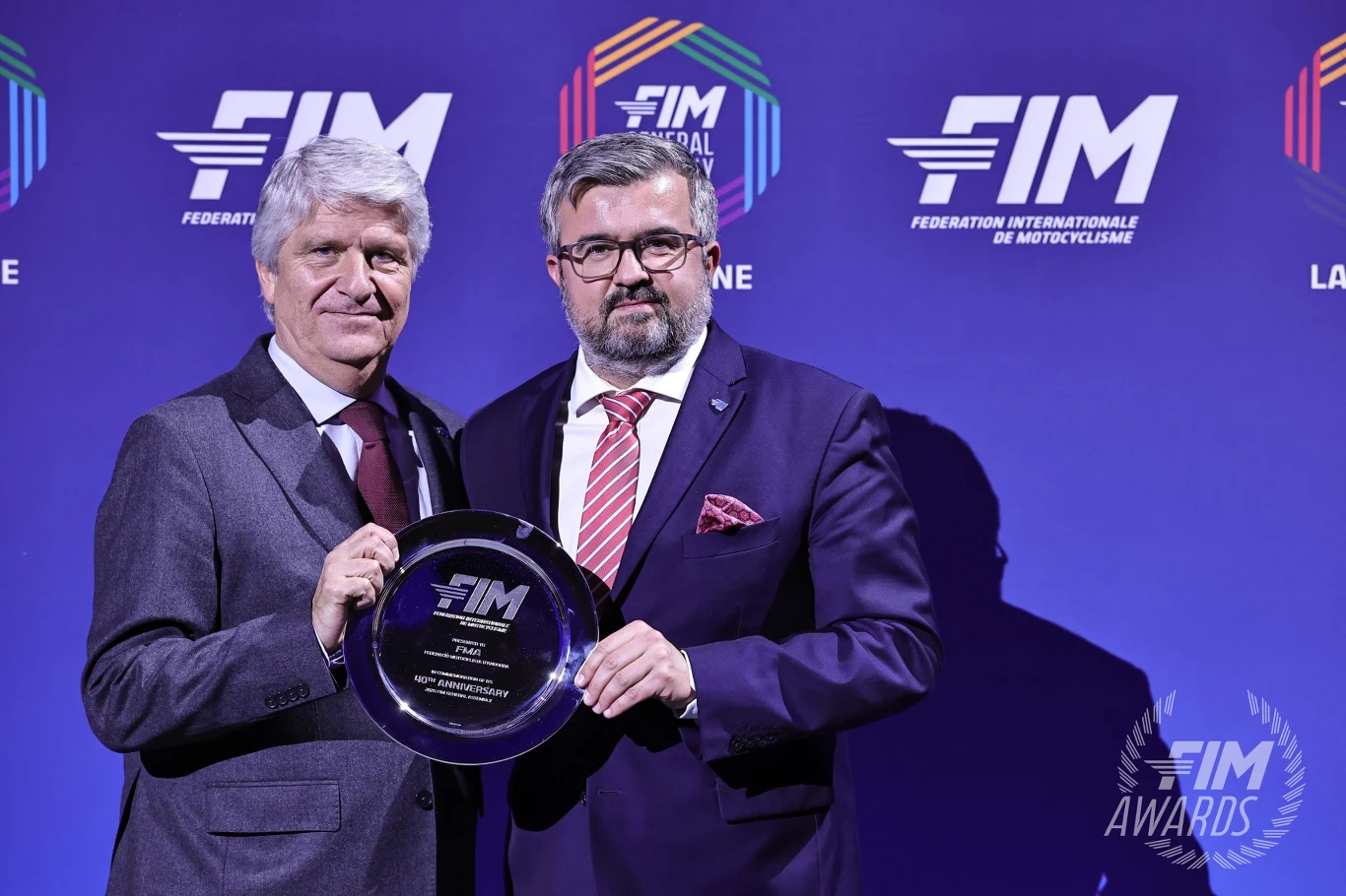 Prezes Michał Sikora z prezydentem FIM Jorge Viegasem. Dwóch elegancko ubranych mężczyzn na tle niebieskiej ścianki z logotypami FIM, jeden z nich trzyma srebrną plakietę z napisem FIM Awards, obaj patrzą prosto w obiektyw.
