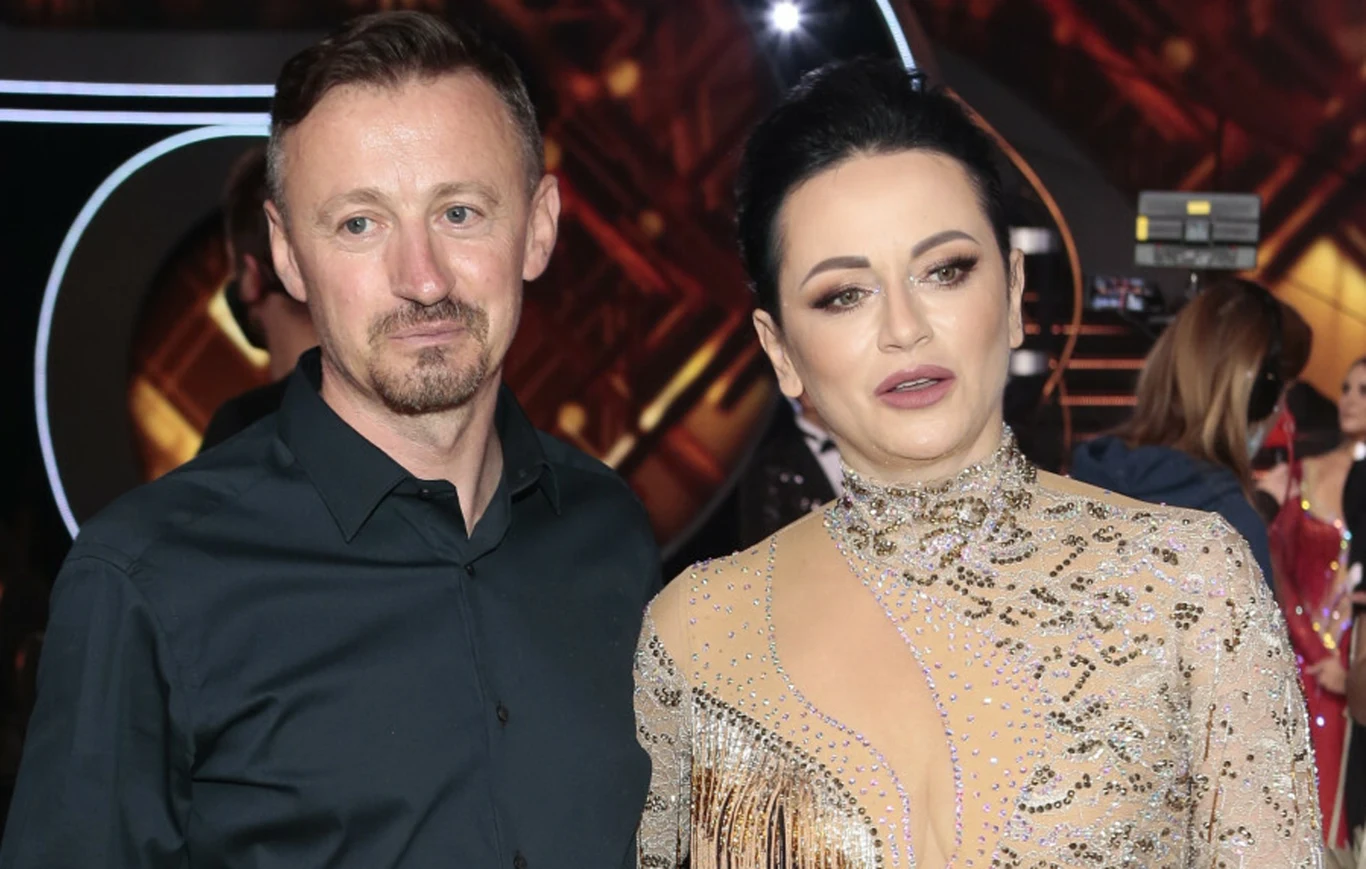 Para elegancko ubranych osób na tle dekoracji scenicznej, kobieta w błyszczącej, beżowej sukni z ozdobami, mężczyzna w ciemnej koszuli, w tle widoczny fragment kamery i światła sceniczne.