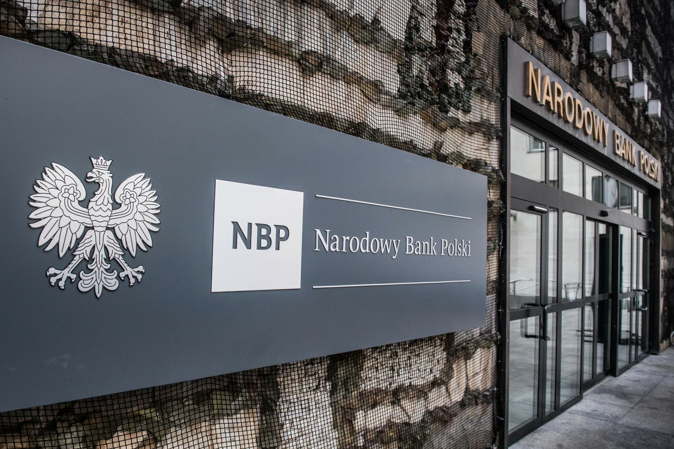 Wejście do siedziby Narodowego Banku Polskiego, duża tablica z logo NBP i godłem Polski zamontowana na elewacji obok szklanych drzwi wejściowych.