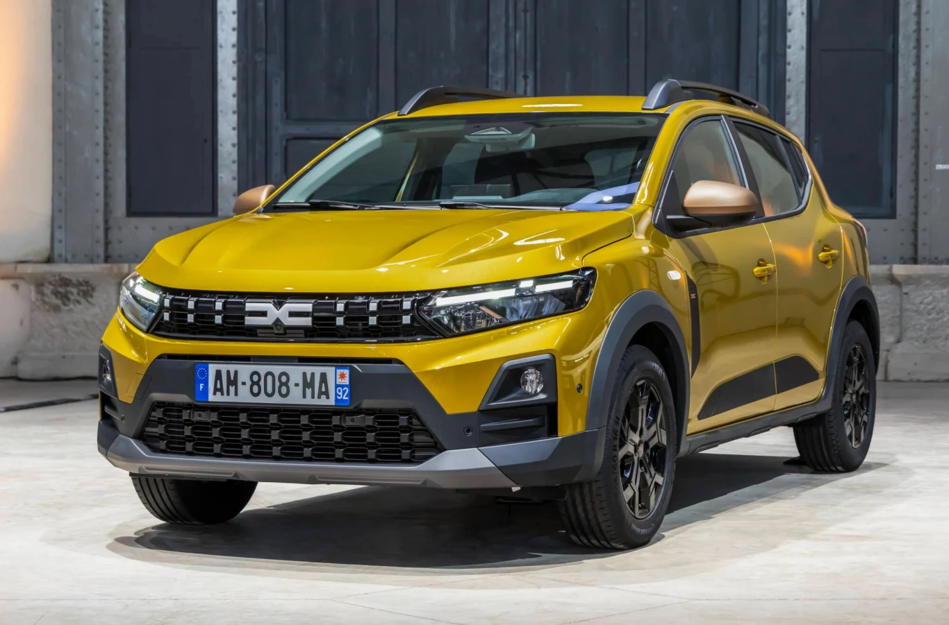 Dacie po liftingu trafiły do polskich salonów. Znamy ceny. Nowoczesny żółty samochód typu SUV stojący w dobrze oświetlonym, industrialnym otoczeniu. Pojazd ma czarne elementy nadwozia, nowoczesne reflektory, czarne felgi i widoczną rejestrację francuską.