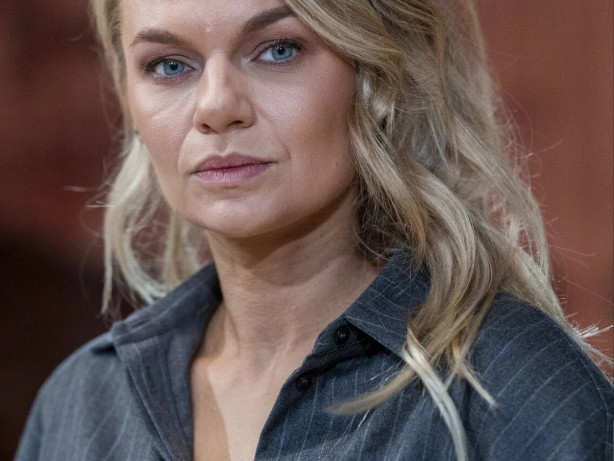 Emilia Komarnicka Blondynka o niebieskich oczach z delikatnym makijażem w szarej koszuli z lekko rozpuszczonymi włosami, patrząca bezpośrednio w obiektyw na tle ciepłych, rozmytych barw.
