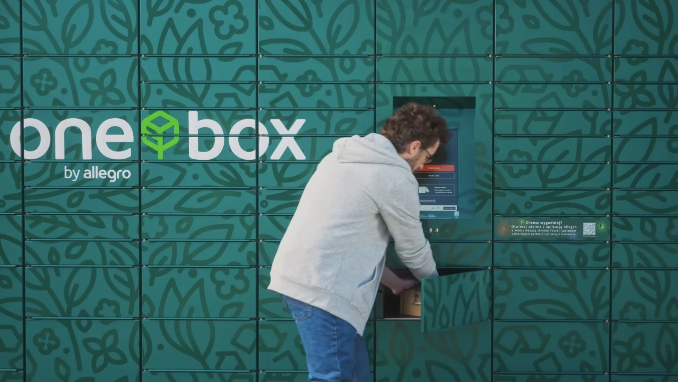 Allegro One Box z nową usługą. Wyślij paczkę do kogo chcesz. Mężczyzna w jasnej bluzy odbiera paczkę z automatu paczkowego na tle zielonych skrytek z napisem one box by allegro.