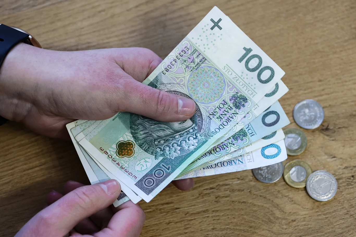 Czy premia jest opodatkowana? Ile pieniędzy trzeba oddać fiskusowi? Ręce trzymające polskie banknoty stuzłotowe oraz drobne monety leżące na drewnianym blacie.
