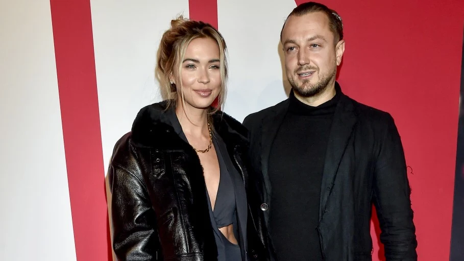 Sandra Kubicka i Baron Dwoje elegancko ubranych ludzi pozuje razem na tle ściany w biało-czerwonych barwach. Kobieta ma jasne włosy spięte do tyłu i nosi ciemną kurtkę oraz sukienkę z głębokim dekoltem. Mężczyzna jest ubrany w czarny garnitur i czarną koszulę.