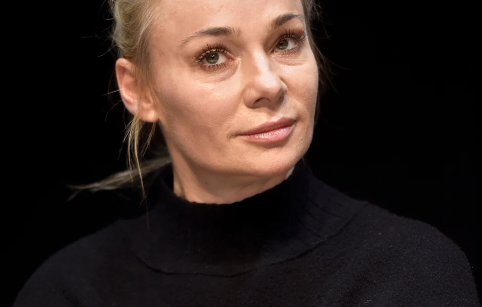 Blondynka o związanych włosach, ubrana w czarny golf, patrzy w kierunku obiektywu na ciemnym tle.