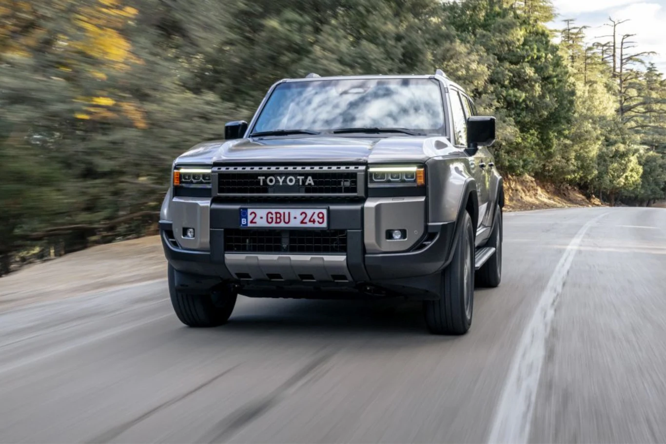 Nowa Toyota Land Cruiser wjeżdża do Polski. Kupisz ją w promocyjnej cenie. Toyota Land Cruiser w nowoczesnej wersji, jadąca pustą asfaltową drogą przez zalesiony teren, samochód z przodu podkreślający masywność, a w tle rozmyte drzewa.