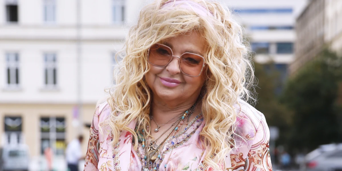 Magda Gessler Kobieta o długich, kręconych blond włosach uśmiecha się delikatnie, nosi duże okulary przeciwsłoneczne, pastelową apaszkę i różowo-beżowy wzorzysty strój, w tle zabudowa miejska z rozmazanym ruchem ulicznym.