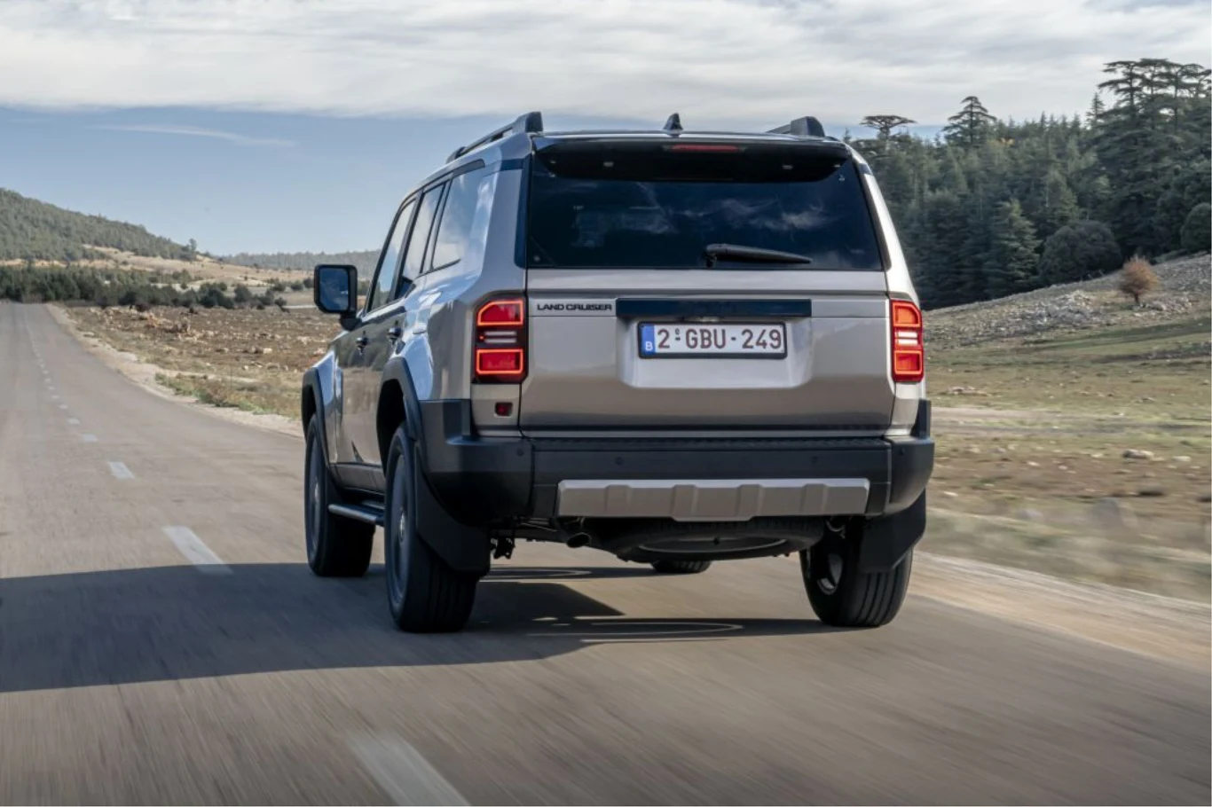 Bagażnik oferuje pojemność 573 l. Pod podłogą ukryta została bateria litowo-jonowa. Samochód terenowy Land Rover Defender jedzie asfaltową drogą przez surowy krajobraz, wokół rozciągają się pola i nieliczne drzewa, na horyzoncie widać góry oraz niebo pokryte lekkimi chmurami.