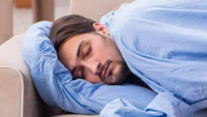 Mężczyzna z brodą śpi na kanapie, na niebieskiej poduszce, przykryty kołdrą