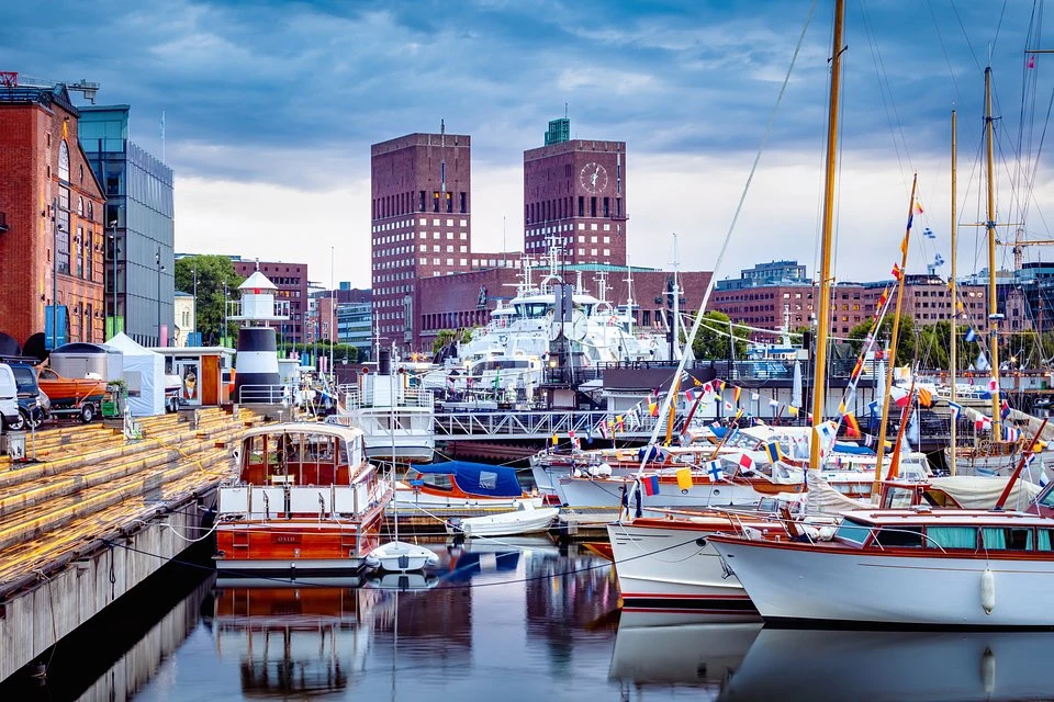 Oslo - esencja skandynawskiej energii. Nowoczesny port jachtowy z licznymi łodziami i jachtami cumującymi przy nabrzeżu, w tle charakterystyczny budynek ratusza z dwoma bliźniaczymi wieżami, po lewej stronie ceglane zabudowania i drewniane schody prowadzące w stronę wody, na niebie widoczne...